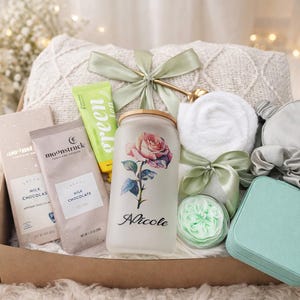 May include: A gift basket filled with self-care items. Includes a blanket, chocolate bars, a jar with a rose design and the name "Nicole", a tube of green lotion, a white washcloth, a sleep mask, a scrunchie, and a small teal box.