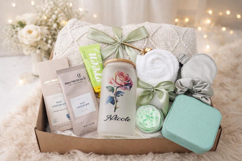 May include: A gift basket filled with self-care items. Includes a blanket, chocolate bars, a jar with a rose design and the name "Nicole", a tube of green lotion, a white washcloth, a sleep mask, a scrunchie, and a small teal box.