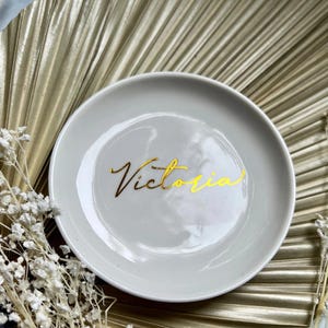 May include: A white ceramic trinket dish with a gold inscription that reads "Victoria".