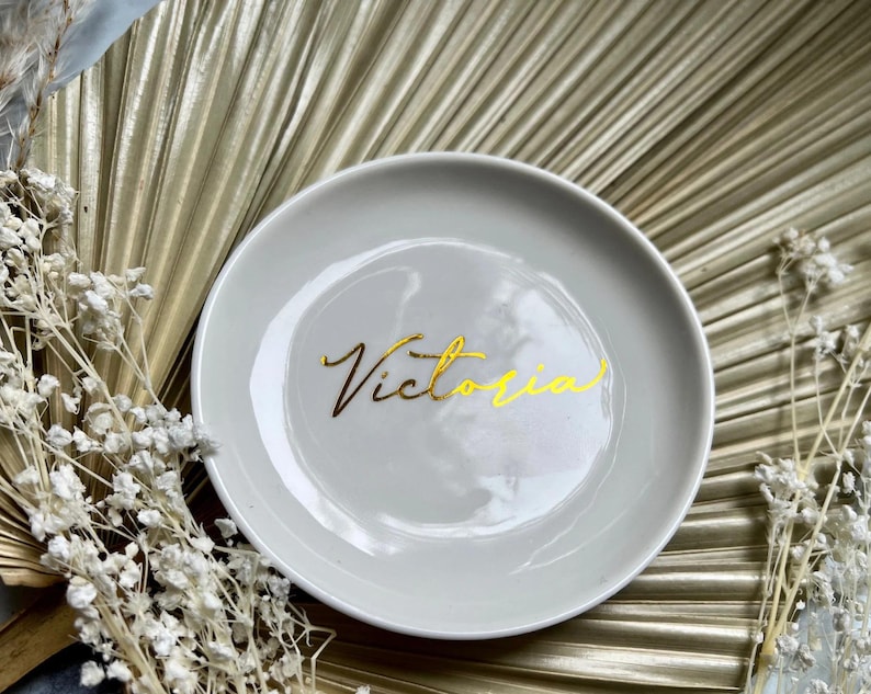May include: A white ceramic trinket dish with a gold inscription that reads "Victoria".
