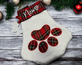 May include: A white Christmas stocking with a red and green plaid paw print design. The cuff is plaid with the name "Nemo" in white script. A white bone-shaped ornament hangs from a red ribbon. Festive holiday decor.