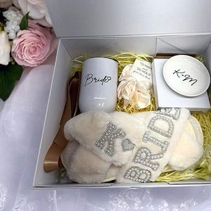 May include: A white gift box filled with bridal shower gifts. The box contains a white mug with the word "Bride" on it, a pair of white fluffy slippers with the word "Bride" in rhinestones, a white dish with the initials "K-M" and a small white card with the text "To have and to hold, your love is gold".