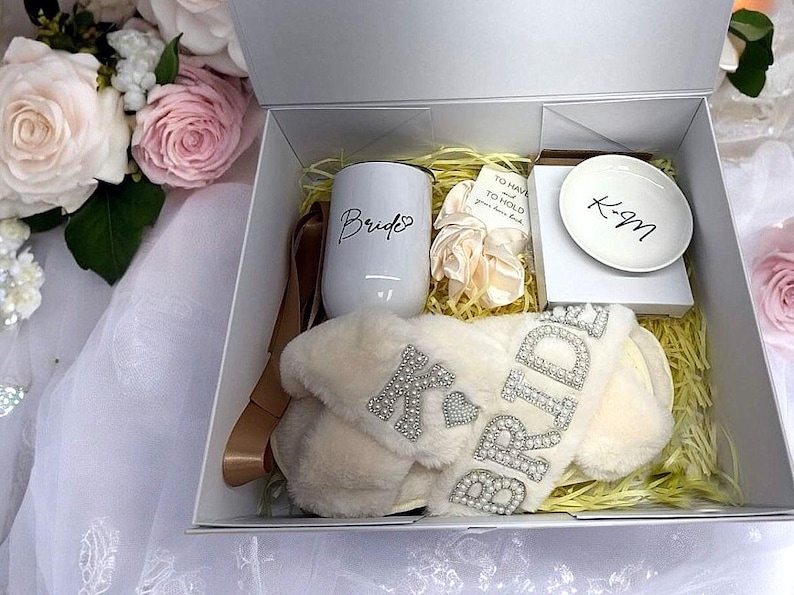 May include: A white gift box filled with bridal shower gifts. The box contains a white mug with the word "Bride" on it, a pair of white fluffy slippers with the word "Bride" in rhinestones, a white dish with the initials "K-M" and a small white card with the text "To have and to hold, your love is gold".