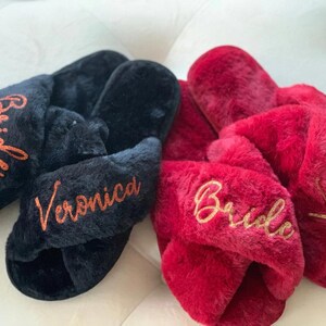 May include: Two pairs of plush, cross-band slippers. One pair is black with "Bride" and "Veronica" embroidered in orange. The other pair is red with "Bride" and "Veronica" embroidered in gold. These slippers are a cozy gift.