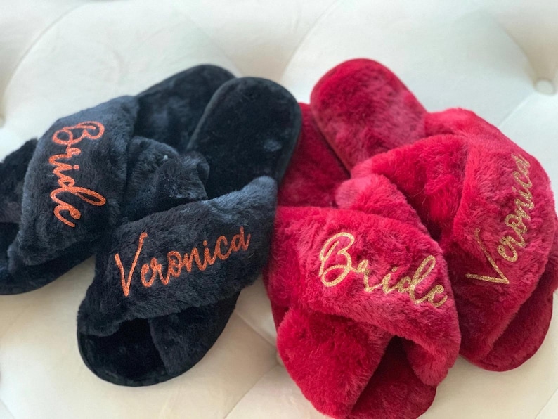 May include: Two pairs of plush, cross-band slippers. One pair is black with "Bride" and "Veronica" embroidered in orange. The other pair is red with "Bride" and "Veronica" embroidered in gold. These slippers are a cozy gift.