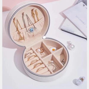 May include: A white, round jewelry box with a zipper, open to reveal various gold and silver necklaces, rings, and earrings. The jewelry box has a light pink interior and is set against a white background. Some jewelry pieces include an evil eye pendant and a pearl earring.