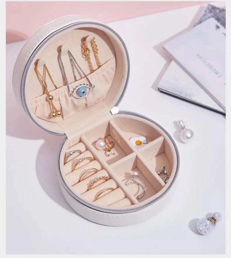 May include: A white, round jewelry box with a zipper, open to reveal various gold and silver necklaces, rings, and earrings. The jewelry box has a light pink interior and is set against a white background. Some jewelry pieces include an evil eye pendant and a pearl earring.