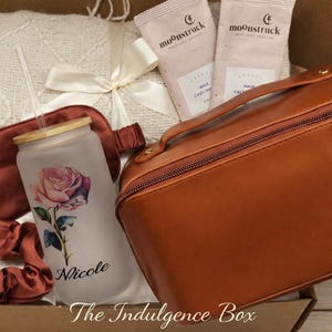 May include: An open gift box containing a variety of items. A frosted glass tumbler with a rose design and the name "Nicole" is visible, along with a brown cosmetic bag, eye mask, scrunchie, and two packets. A white blanket and a bow are also included.