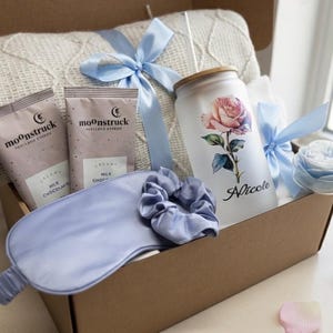 May include: A gift box filled with a white knit blanket, two packages of milk chocolate, a frosted glass tumbler with a rose design and the name "Nicole", a blue sleep mask, a scrunchie, and a blue ribbon.