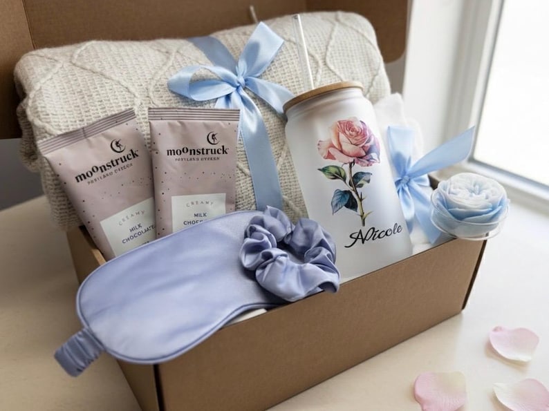 May include: A gift box filled with a white knit blanket, two packages of milk chocolate, a frosted glass tumbler with a rose design and the name "Nicole", a blue sleep mask, a scrunchie, and a blue ribbon.