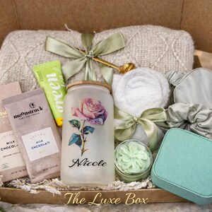 May include: A gift box filled with self-care items. Includes a floral-decorated glass, chocolate bars, a green tube, a small jar of cream, a towel, an eye mask, a scrunchie, and a teal square box. The box is labeled "The Luxe Box".