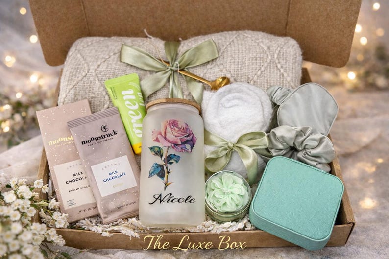 May include: A gift box filled with self-care items. Includes a floral-decorated glass, chocolate bars, a green tube, a small jar of cream, a towel, an eye mask, a scrunchie, and a teal square box. The box is labeled "The Luxe Box".