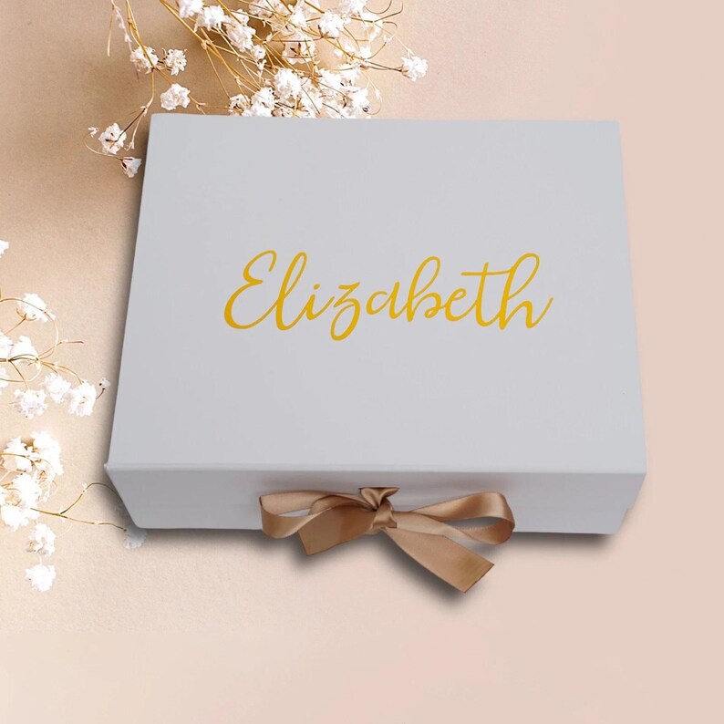 May include: A white gift box with the name "Elizabeth" in gold script on the lid. A gold ribbon is tied in a bow at the front. The box is on a light pink surface, with white flowers in the background.