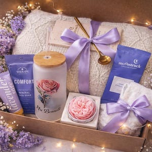 May include: A gift box filled with self-care items, including a glass with a rose design, a bar of soap, a small towel, and a chocolate bar. The box is tied with a lavender ribbon and surrounded by purple flowers.