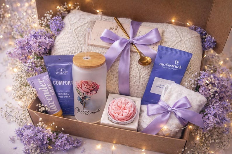 May include: A gift box filled with self-care items, including a glass with a rose design, a bar of soap, a small towel, and a chocolate bar. The box is tied with a lavender ribbon and surrounded by purple flowers.
