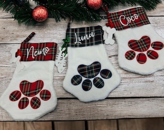 May include: Three white Christmas stockings with a paw print design. Each stocking has a red and green plaid cuff with a name embroidered in white. Bone-shaped ornaments hang from the stockings. Festive holiday decor.