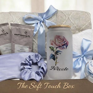 May include: A gift box with a variety of self-care items. Includes a blanket, two packets of milk chocolate, a sleep mask, a scrunchie, a glass with a rose design, a towel, and a decorative item. The box is tied with light blue ribbons. Text on the glass reads "Nicole".