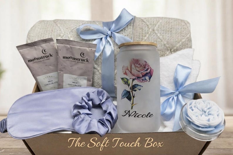 May include: A gift box with a variety of self-care items. Includes a blanket, two packets of milk chocolate, a sleep mask, a scrunchie, a glass with a rose design, a towel, and a decorative item. The box is tied with light blue ribbons. Text on the glass reads "Nicole".