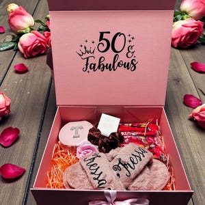 May include: A pink gift box with the text "50 & Fabulous" contains a variety of items. Inside are pink slippers with the name "Tressa", a compact, hair accessories, and a floral face mask. Pink roses and petals surround the box.