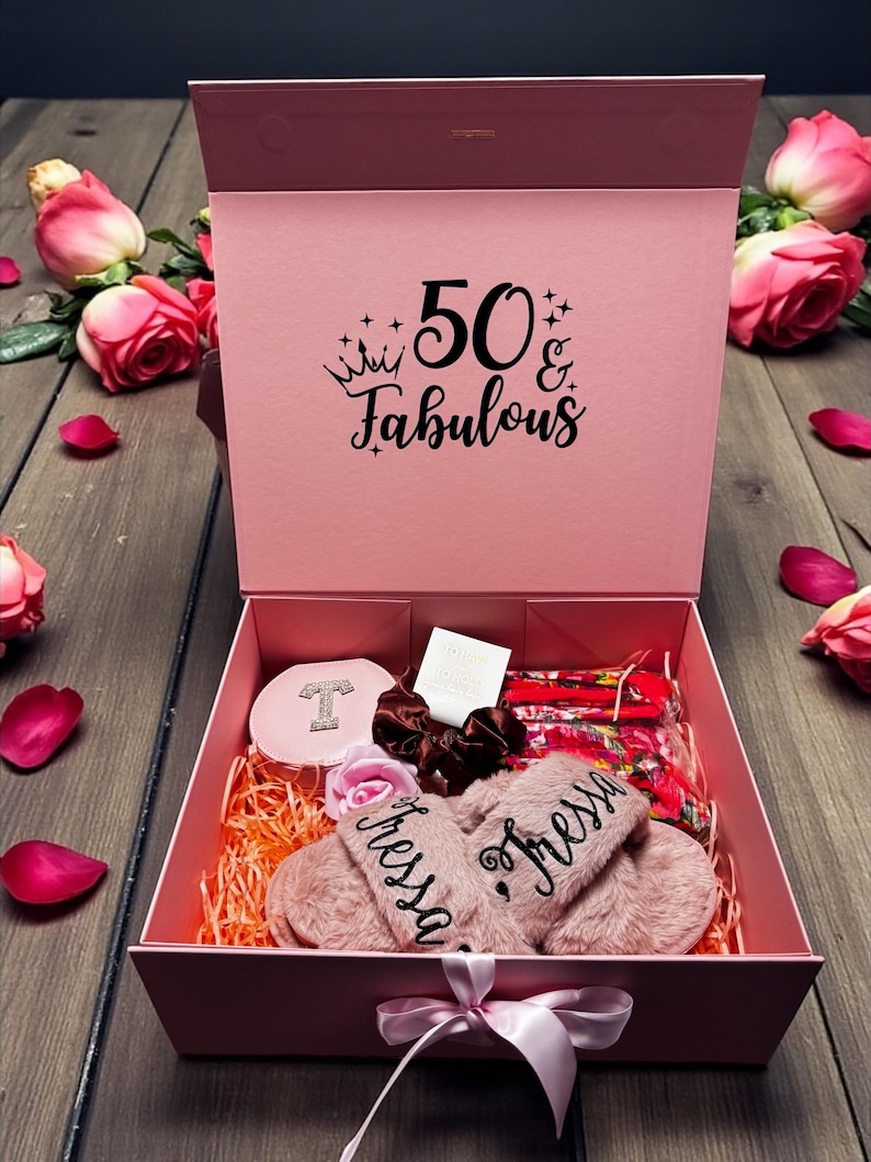 May include: A pink gift box with the text "50 & Fabulous" contains a variety of items. Inside are pink slippers with the name "Tressa", a compact, hair accessories, and a floral face mask. Pink roses and petals surround the box.
