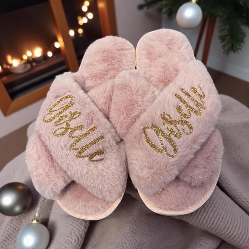 May include: A pair of light pink, fluffy cross-band slippers. The word "Giselle" is embroidered in gold glitter across the top of each slipper. The slippers are resting on a gray blanket, with a blurred background of Christmas decorations.