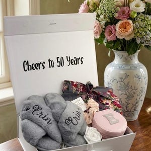 May include: A white gift box with the text "Cheers to 50 Years" contains gray slippers embroidered with "Erin", a floral print robe, a pink jewelry case, and a white rose. A bouquet of flowers sits nearby.