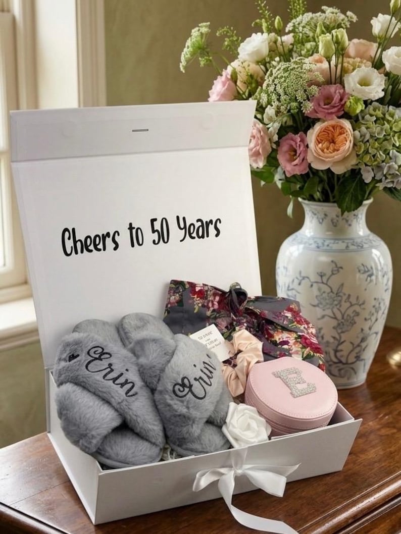 May include: A white gift box with the text "Cheers to 50 Years" contains gray slippers embroidered with "Erin", a floral print robe, a pink jewelry case, and a white rose. A bouquet of flowers sits nearby.