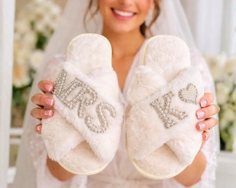 May include: Fluffy white cross-band slippers with the word "MRS" spelled out in silver rhinestones on one slipper and the letter "K" and a heart on the other. The slippers are a bridal accessory.