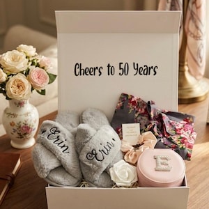 May include: A gift box with the text "Cheers to 50 Years" contains gray slippers embroidered with "Erin", a floral robe, scrunchies, a jewelry case with the letter "E", and a white rose. A vase of flowers is on the left.