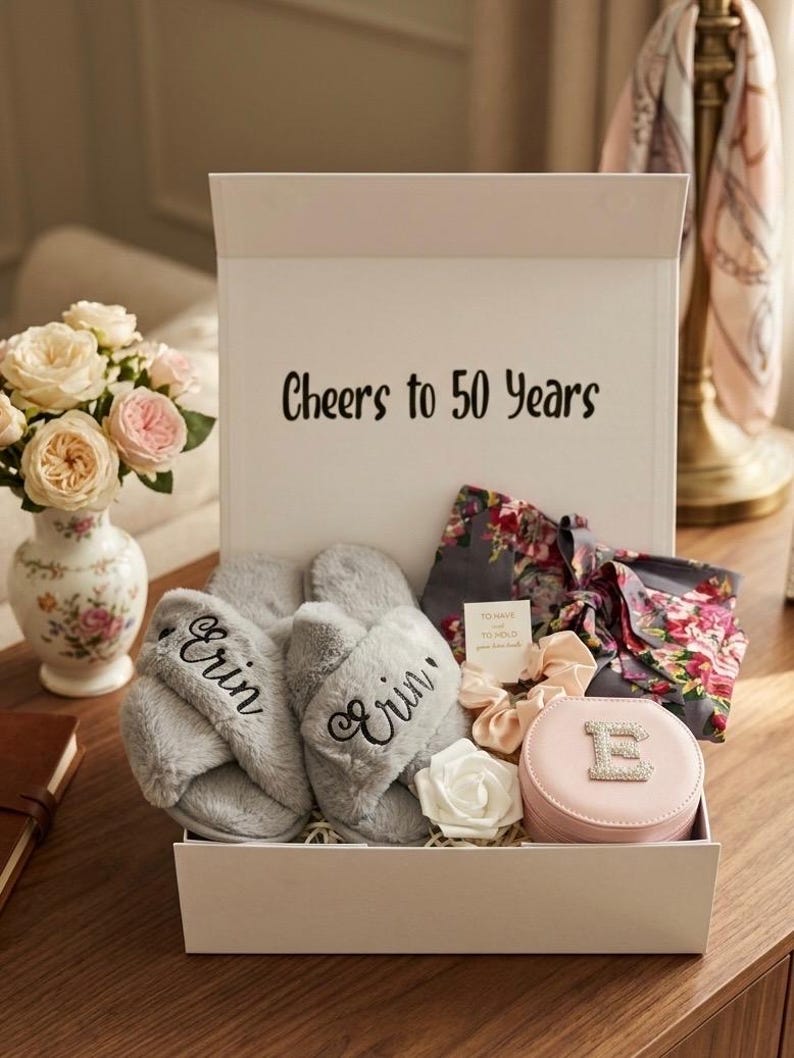May include: A gift box with the text "Cheers to 50 Years" contains gray slippers embroidered with "Erin", a floral robe, scrunchies, a jewelry case with the letter "E", and a white rose. A vase of flowers is on the left.