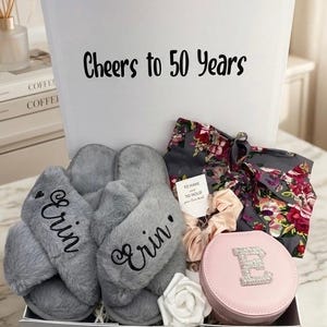 May include: A gift box with the text "Cheers to 50 Years" contains gray slippers embroidered with "Erin", a floral robe, a pink jewelry case with a rhinestone "E", and other accessories. A white rose and scrunchies are also included.