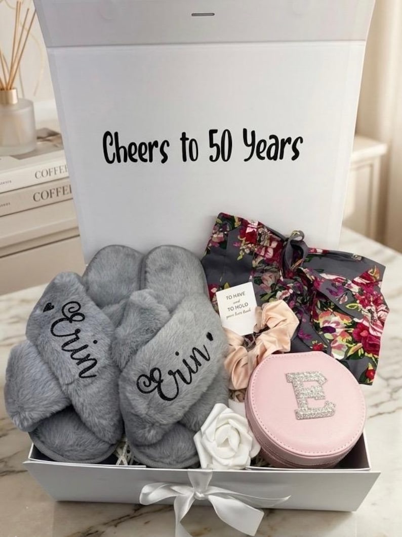 May include: A gift box with the text "Cheers to 50 Years" contains gray slippers embroidered with "Erin", a floral robe, a pink jewelry case with a rhinestone "E", and other accessories. A white rose and scrunchies are also included.