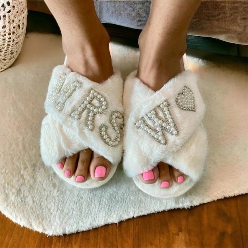 May include: White fluffy slippers with a criss-cross strap design. The slippers have pearl embellishments spelling out "MRS" and "W" with a heart on one slipper.