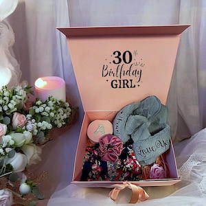 May include: A pink gift box with a lid that says "30th Birthday Girl" is open to reveal a gift set. The set includes a pair of gray fuzzy slippers, a small round box with a gold letter "A" on it, a pink hair scrunchie, and a floral patterned fabric.