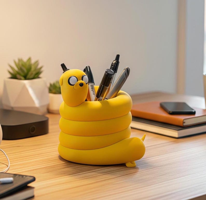 May include: A yellow, cylindrical desk organizer shaped like a coiled cartoon dog character with large white eyes and a black nose. The hollow center holds several black and silver pens and markers, with the dog's head extending from the top rim.
