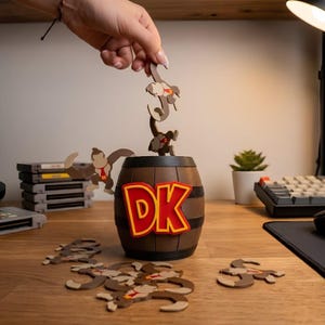 May include: A brown barrel with the letters "DK" in red and yellow, surrounded by wooden monkey cutouts. A hand is holding a monkey cutout above the barrel. Several game cartridges are in the background.