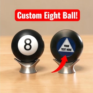 May include: Two black spherical eight balls resting on silver metal stands. The left ball features a white circle with a black number eight. The right ball displays a blue triangle with the text YOUR TEXT HERE and a red arrow pointing upward.