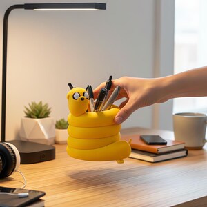May include: A bright yellow pencil holder shaped like the cartoon character Jake the Dog from Adventure Time. The cylindrical container features a smooth, coiled body design with a sculpted head at the top, currently holding several black and silver pens.
