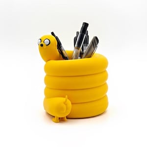 May include: A bright yellow cylindrical desk organizer shaped like a cartoon dog with a coiled body. The character has a rounded head with two large white eyes and a small black nose. Several black and silver pens are stored inside the container.