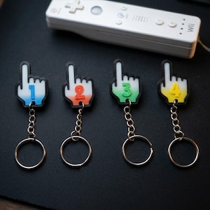May include: Four keychains shaped like pointing fingers, each featuring a different number from one to four. The first is blue, the second is orange, the third is green, and the fourth is yellow. Each keychain has a metal chain and ring.