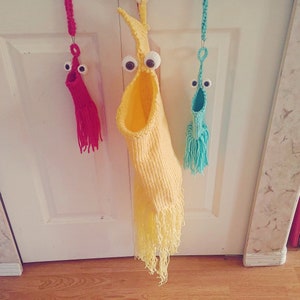 Puede incluir: Tres adornos de calcetines de punto con ojos saltones. Los adornos son amarillos, rosas y azules. El adorno amarillo tiene una franja en la parte inferior.