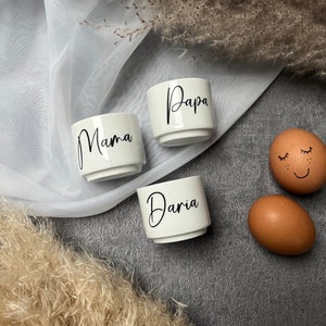 Può includere: Tre portauova in ceramica bianca con le parole "Mama", "Papa" e "Daria" in corsivo nero. Due uova marroni sono accanto alle tazze, una con un volto disegnato. Le tazze sono su una superficie grigia con piante secche decorative.