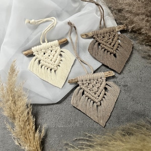Puede incluir: Tres tapices de macramé de pared con forma triangular y flecos. Los tapices están hechos con fibras naturales y tienen un estilo rústico y bohemio. Están colgados de un fondo de tela blanca transparente.