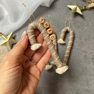 Könnte beinhalten: Zwei beige-weiße Seil-Zuckerstangen-Ornamente mit Holzperlen, die den Namen "Theo" buchstabieren.