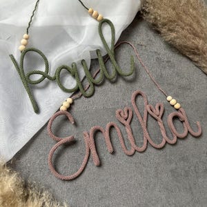 Op de afbeelding: Twee wandhangende naambordjes gemaakt van gebreid garen. Het bovenste bordje is olijfgroen en spelt de naam Paul. Het onderste bordje is oudroze en spelt de naam Emilia, met twee hartjes in de letters verwerkt. Beide hebben houten kralen.