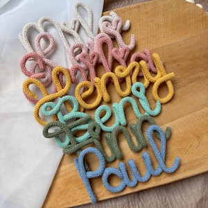 Könnte beinhalten: Eine Sammlung personalisierter Namen aus gestricktem Garn in verschiedenen Farben wie Creme, Rosa, Gelb, Türkis, Olivgrün und Hellblau. Jeder Name ist in einer durchgehenden Schreibschrift geformt und weist weiche, strukturierte Stoffschlingen auf.