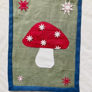 Op de afbeelding: Een handgemaakt textielkunstwerk met een rode paddenstoel en witte sterren op een groene achtergrond. Het stuk is omlijst met een blauwe rand en bevat stervormige versieringen. Het kunstwerk is waarschijnlijk een decoratief item.