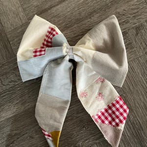 May include: A large, handmade fabric bow tie with a patchwork design. The bow tie incorporates various fabrics, including red gingham, floral prints, and striped patterns. The colour palette includes cream, red, blue, and tan.