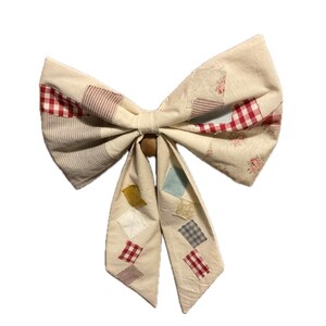 Puede incluir: Un lazo grande de tela color crema con un diseño de patchwork. El lazo presenta cuadrados de cuadros rojos, estampados florales y a rayas. El centro del lazo está atado con una cuenta de madera, y las colas tienen parches en forma de diamante.