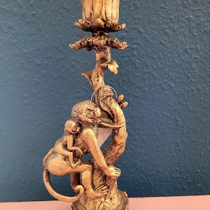 May include: A gold-colored candle holder featuring a monkey and baby monkey climbing a tree-like structure. The top of the holder has a flower-like design to hold a candle. The base is ornate and detailed.